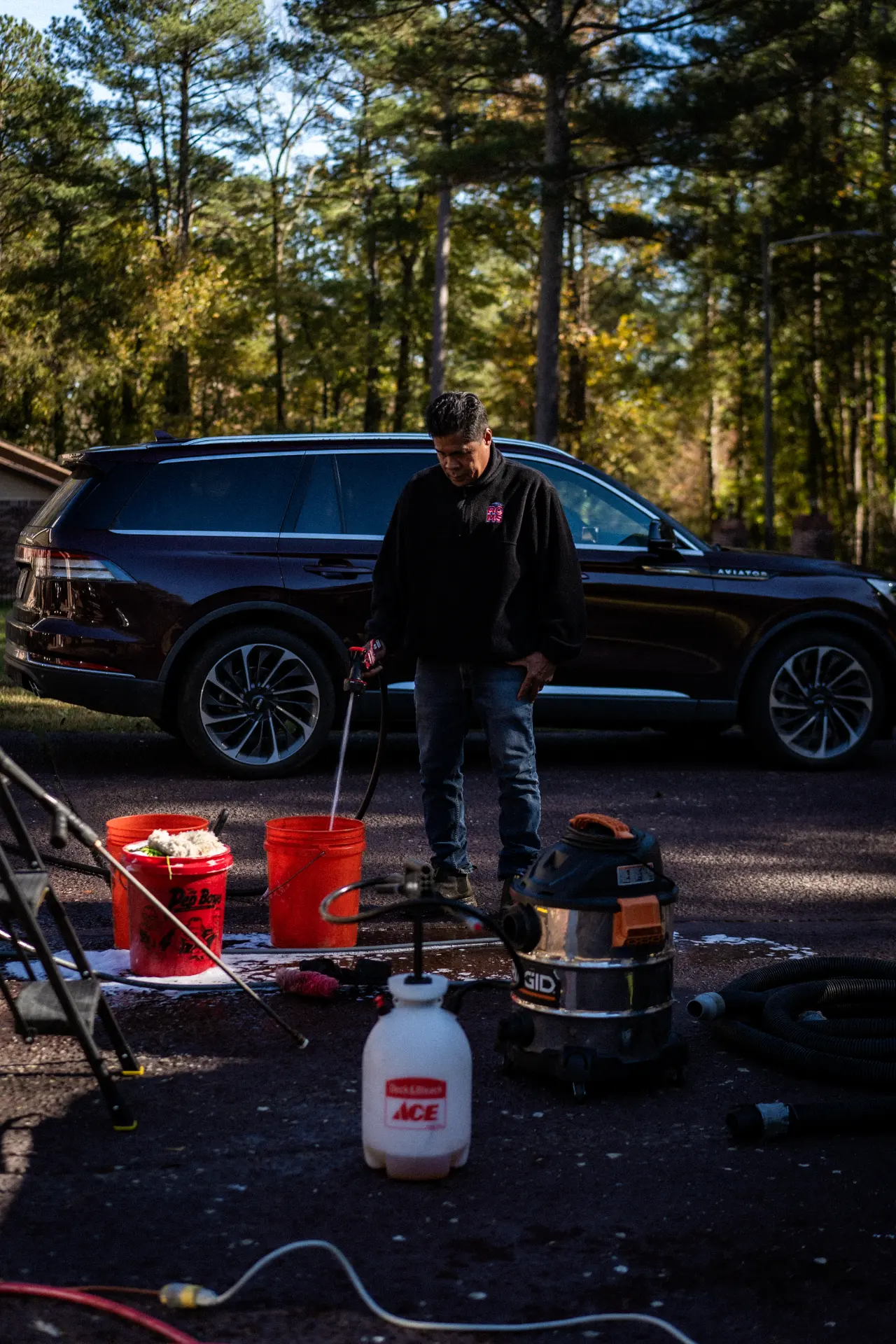 Peter with buckets and a pump sprayer beside a dark Lincoln Aviator on a trailer